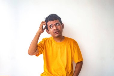 Sarı gömlekli bir adam, kafası karışık bir ifadeyle kaşınıyor, düz beyaz bir arkaplanda oturuyor. Bu görüntü bir kafa karışıklığı ya da belirsizlik anını yakalar