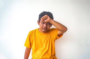 Sarı gömlekli bir adam, tek eliyle başını tutarak düz beyaz bir zemine yaslanır. Yorgunluk ve yorgunluk belirtisi gösterir. Görüntü yorgunluk anını yakalar.