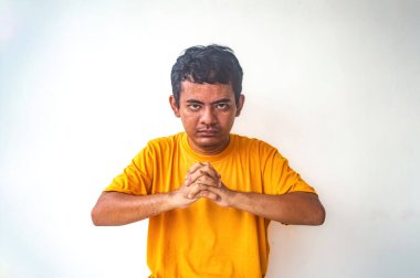 Sarı gömlekli bir adam düz beyaz bir arkaplana karşı duruyor. Elleri önünde birbirine kenetlenmiş, ciddi ve yoğun bir jest yapıyor.