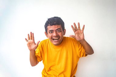 Sarı tişörtlü bir adam iki elini de açık bir şekilde kaldırıyor. Beyaz arka plan karşısında şaşırmış ya da ürkmüş gibi görünüyor.