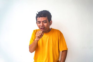 Sarı gömlekli bir adam elleriyle öksürüğe benzeyen bir jest yapıyor. Düz beyaz arkaplan jesti vurgular, onu görüntünün odak noktası yapar.