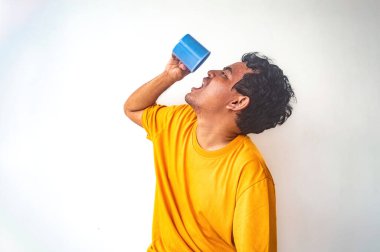 Sarı gömlekli bir adam mavi bir bardaktan su içiyor, sanki çok susamış gibi başını arkaya eğiyor. Düz beyaz arka plan onun jestini vurguluyor.