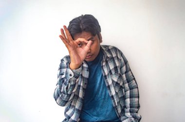 Ekose gömlekli ve mavi tişörtlü bir adam üç parmağı havada, düz beyaz bir arkaplana karşı duran iyi bir el hareketi yapıyor.