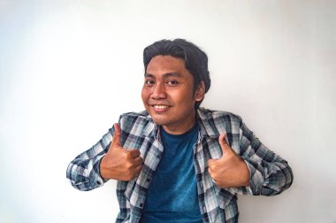 Ekose gömlekli ve mavi tişörtlü bir adam, beyaz bir arka plana karşı yüzünde kocaman bir gülümsemeyle çifte baş parmak işareti yapıyor. Bu görüntü bir neşe ve onay anı yakalar.