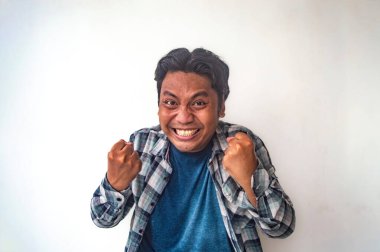 Ekose gömlekli ve mavi tişörtlü bir adam düz beyaz bir arkaplan önünde iki elini de sıkıyor, bir başarı veya başarı jesti sergiliyor. Bu görüntü zafer ve neşe dolu bir anı yakalar.