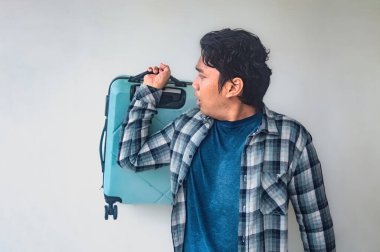 Ekose gömlekli ve mavi tişörtlü bir adam elinde mavi bir bavul tutuyor. Merakını ya da şaşkınlığını ifade ediyor. Düz beyaz arka plan, bir seyahat teması yakalayarak hareketi vurguluyor.