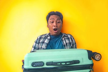 Ekose gömlekli ve mavi tişörtlü bir adam elinde yeşil bir bavul tutarken abartılı bir sürpriz jest yapıyor. Parlak sarı bir arkaplanda izole edilmiş, aşırı şok anını yakalıyor.