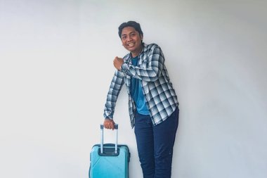 Ekose gömlekli ve mavi pantolonlu bir adam beyaz bir duvara karşı duruyor, elinde uzun saplı mavi bir bavul var. Adam tek kolla hareket ediyor, seyahatin heyecanını yakalıyor.