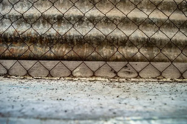 Tel örgüler ve yıpranmış ahşap kirişler paslanmış metal levhaların arka planına kurulmuş desenli bir sınır oluşturuyor. Bu resim ham maddeleri ve kentsel bozulmayı vurgular