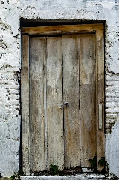 Kırsal, yıpranmış ahşap bir kapı. Parçalanmış, beyaz, yıkanmış tuğla bir duvarda sergilenen dokular, çürüme ve unutulmuş bir tarih hissi.