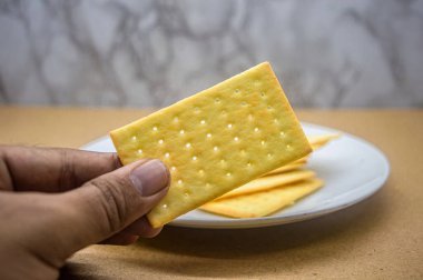 Bir el ön planda tek bir altın, dikdörtgen kraker tutar, yumuşak odaklı arka planda beyaz bir tabakta bir yığın vardır. Atıştırmalık ya da yemek fotoğrafçılığı için ideal.
