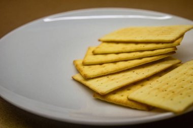 Beyaz bir tabakta altın, dikdörtgen kraker yığını, yumuşak odaklı arka plan. Yemek sunumları, abur cubur fikirleri ya da mutfak blogları için mükemmel.