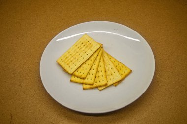 Beyaz bir tabakta, kahverengi arka planda duran bir yığın gevrek, dikdörtgen kraker. Atıştırmalık, yemek ve aperatif temaları için ideal