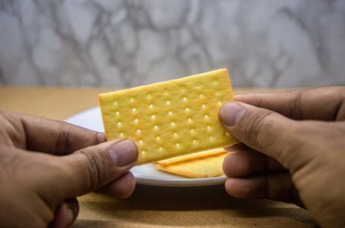 İki el tek bir altın, dikdörtgen kraker tutuyor, yenmeye hazır. Arka planda bir tabak daha kraker yavaşça bulanıklaşıyor. Atıştırmalık veya yemek temaları için ideal