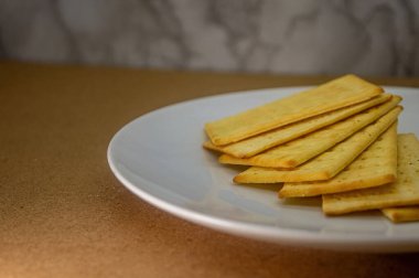 Beyaz bir plaka üzerinde altın, dikdörtgen krakerler yığını, kahverengi bir yüzeye ayarlanmış ve ince dokulu bir arka plan. Gıda içeriği için ideal