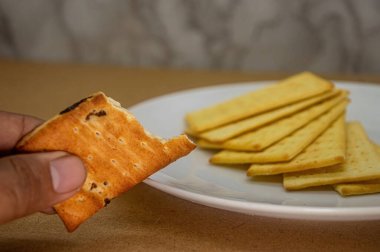 Bir el ısırılmış altın sarısı, dikdörtgen bir kraker tutar, ve arka planda beyaz bir tabakta bir yığın kraker bulunur. Atıştırmalık ya da yemek fotoğrafçılığı için ideal.
