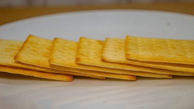 Beyaz bir tabakta düzgün bir sıraya dizilmiş birkaç altın, dikdörtgen krakerin yakın bir yan görüntüsü. Gıda reklamları veya atıştırmalık içeriği için ideal