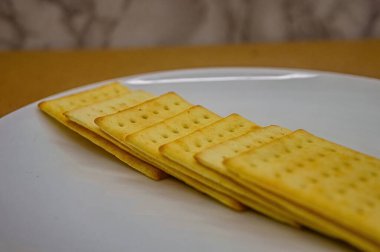 Beyaz bir tabakta düzgün bir sıraya dizilmiş birkaç altın, dikdörtgen krakerin yakın bir yan görüntüsü. Gıda reklamları veya atıştırmalık içeriği için ideal