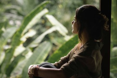 Pencere kenarında oturan Kadın Tropik Doğa 'ya Bakıyor