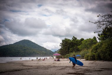 Itamambuca Sahili 'nde Bulutlu bir günde Mavi Sörf Tahtası Taşıyan Adam - Ubatuba, Sao Paulo, Brezilya