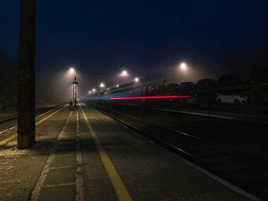Geceleri boş tren istasyonu platformu, arka planda hafif raylarla uzun pozlama, araba nakliye treni.