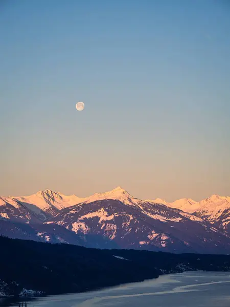 Winter dawn landscape over the Millstatter See in Carinthia, Austria, full moon at sunrise, snow covered mountains, forest, lake