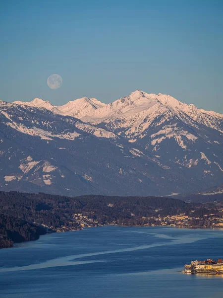 Winter dawn landscape over the Millstatter See in Carinthia, Austria, full moon at sunrise, snow covered mountains, forest, lake