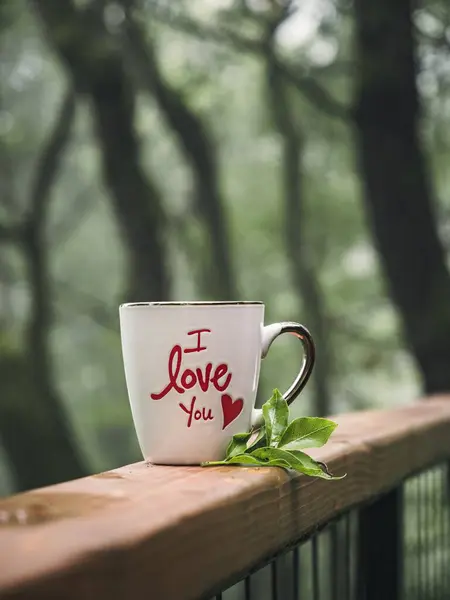 White coffee tea mug with \'I live you\' inscription with tea leaves coffee leaves on a forest balcony porch in the morning, summer nature getaway vacation