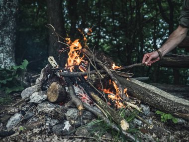 Elleriyle beslenen bir adam orman kamp ateşine odun koyuyor, yüzü yok, boş zamanlarında kamp kuruyor.