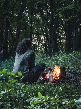 Ormandaki bir kamp ateşinde oturan uzun saçlı insan, arkadan, ateşe bakıyor, yeşil yapraklar yaz, gün ışığı.
