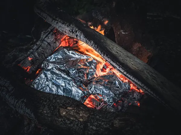 Tin foil packages of grilled food in a campfire, firewood logs ash amber flame, night, dark