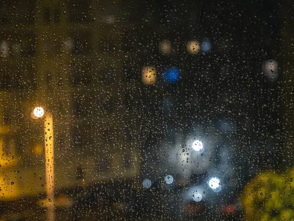 Raindrops on a window during night with colorful blurry city lights street lamps on the outside, warm colors, moody atmosphere