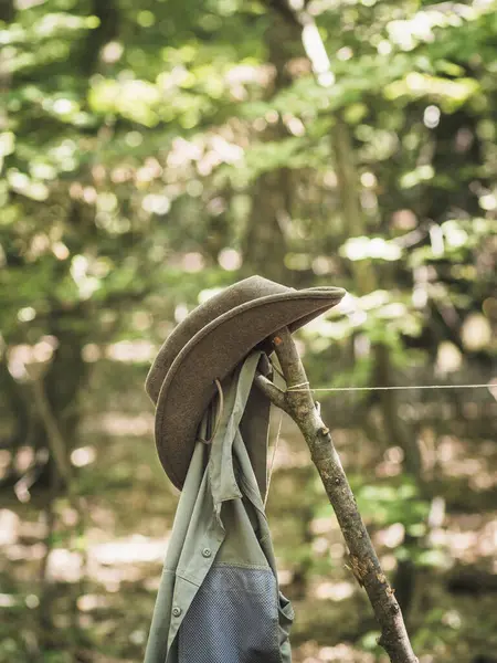Outdoor hiking clothes green olive hat and shirt hanging on a stick in a camp, hunting equipment, nature expedition, camping lifestyle in a forest