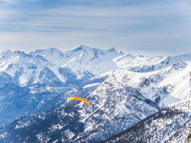 Canlı turuncu kanatlı bir paraglider Fransa 'nın Briancon kentindeki Serre Chevalier' de karla kaplı dağların üzerinden süzülüyor. Parlak gökyüzü ve engebeli tepeler çarpıcı bir dağ manzarası yaratır..