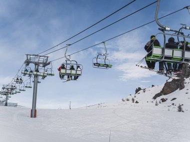 Kayakçılar Briancon, Fransa 'da karlı bir yamaçta, açık bir gökyüzü ve arka planda engebeli bir dağ arazisi olan bir teleferiğe biniyorlar. Sahne kış sporlarının ve dağlık arazilerin özünü yansıtıyor..