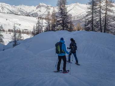 Kar ayakkabılı iki yürüyüşçü, Briancon 'da Serre Chevalier' de derin karların içinde yürürler. Karla kaplı ağaçlar ve görkemli dağ zirveleriyle çevrili bir kış macerasının tadını çıkarıyorlar..
