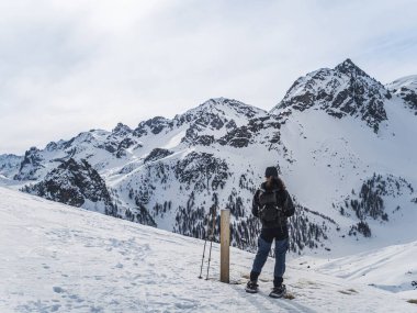 Kar ayakkabıları içinde yalnız bir yürüyüşçü ağaçlarla ve engebeli tepelerle çevrili karlı bir dağ yolunda yürüyor ve doğada huzurlu bir kış macerasının tadını çıkarıyor..