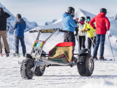 Serre Chevalier 'de açık bir kış gününde arka planda bir grup kayakçıyla birlikte karlı bir yamaca parkedilmiş bir dağ arabası.