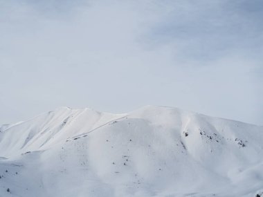 Karlı bir dağ zirvesi bulutlu gri bir gökyüzünün altında uzun duruyor. Yumuşak, el değmemiş yamaçlar ve sessiz tonlar sakin ama dramatik bir alp sahnesi yaratır..