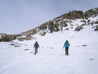 Kar ayakkabısı giyen iki yürüyüşçü Briancon, Fransa 'da Mont Chaberton yakınlarında karlı bir yamaçtan tırmanıyor. Tırmanma direkleriyle donatılmış bulutlu bir gökyüzünün altındaki dağlık arazide yol alırlar..