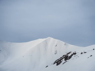 Karlı bir dağ zirvesi bulutlu gri bir gökyüzünün altında uzun duruyor. Yumuşak, el değmemiş yamaçlar ve sessiz tonlar sakin ama dramatik bir alp sahnesi yaratır..