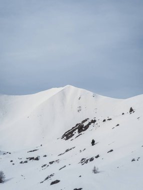 Karlı bir dağ zirvesi bulutlu gri bir gökyüzünün altında uzun duruyor. Yumuşak, el değmemiş yamaçlar ve sessiz tonlar sakin ama dramatik bir alp sahnesi yaratır..