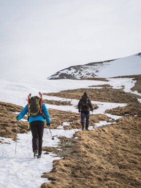 Two hikers with backpacks and trekking poles climb the snowy slopes of Mont Chaberton in France. The rugged terrain blends snow and dry grass under an overcast sky. clipart