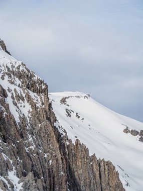 A striking view of a snow-covered mountain ridge with rugged rock formations under a dramatic cloudy sky. The scene captures the raw beauty of the French Alps. clipart