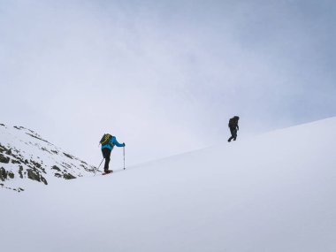 Kar ayakkabısı giyen iki yürüyüşçü Briancon, Fransa 'da Mont Chaberton yakınlarında karlı bir yamaçtan tırmanıyor. Tırmanma direkleriyle donatılmış bulutlu bir gökyüzünün altındaki dağlık arazide yol alırlar..