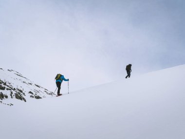 Two hikers wearing snowshoes ascend a snowy slope near Mont Chaberton in Briancon, France. Equipped with trekking poles, they navigate the alpine terrain under a cloudy sky. clipart