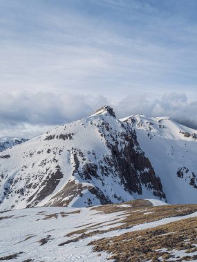 Çarpıcı, karla kaplı bir dağ, bulutları dağınık, berrak, mavi bir gökyüzünün zeminine doğru yükseliyor. Kayalık arazi ve kar parçaları dramatik ve engebeli bir dağ manzarası yaratır..