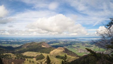 Chiemsee ve Bavyera kırsalının nefes kesici panoramik manzarası. Yuvarlanan tepeler, yemyeşil ormanlar ve büyüleyici köyler dağınık bulutlarla dolu canlı mavi bir gökyüzü altında uzanıyor..