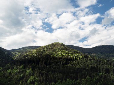 Yoğun yeşil ormanlarla kaplı, parçalı bulutlu bir gökyüzünün altında nefes kesici bir dağ manzarası. Güneş ışığı, ağaçların tepelerini vurgular ve doğal ortamda ışık ile gölge arasında bir zıtlık yaratır..