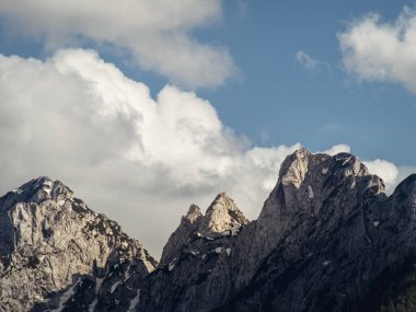 Karla kaplı dramatik kayalık dağ zirveleri bulutlu bir gökyüzüne karşı kurulur. Engebeli arazi ve yüksek uçurumlar yukarıdaki yumuşak bulutlarla çarpıcı bir zıtlık yaratır..